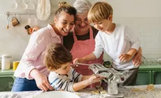 Une famille avec deux jeunes garçons qui font des pâtes ensemble dans la cuisine, guidés par leur mère et leur grand-mère.